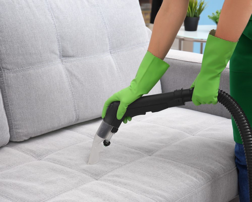 Attractive cleaner in uniform and rubber gloves cleaning upholstery of couch with vacuum cleaner